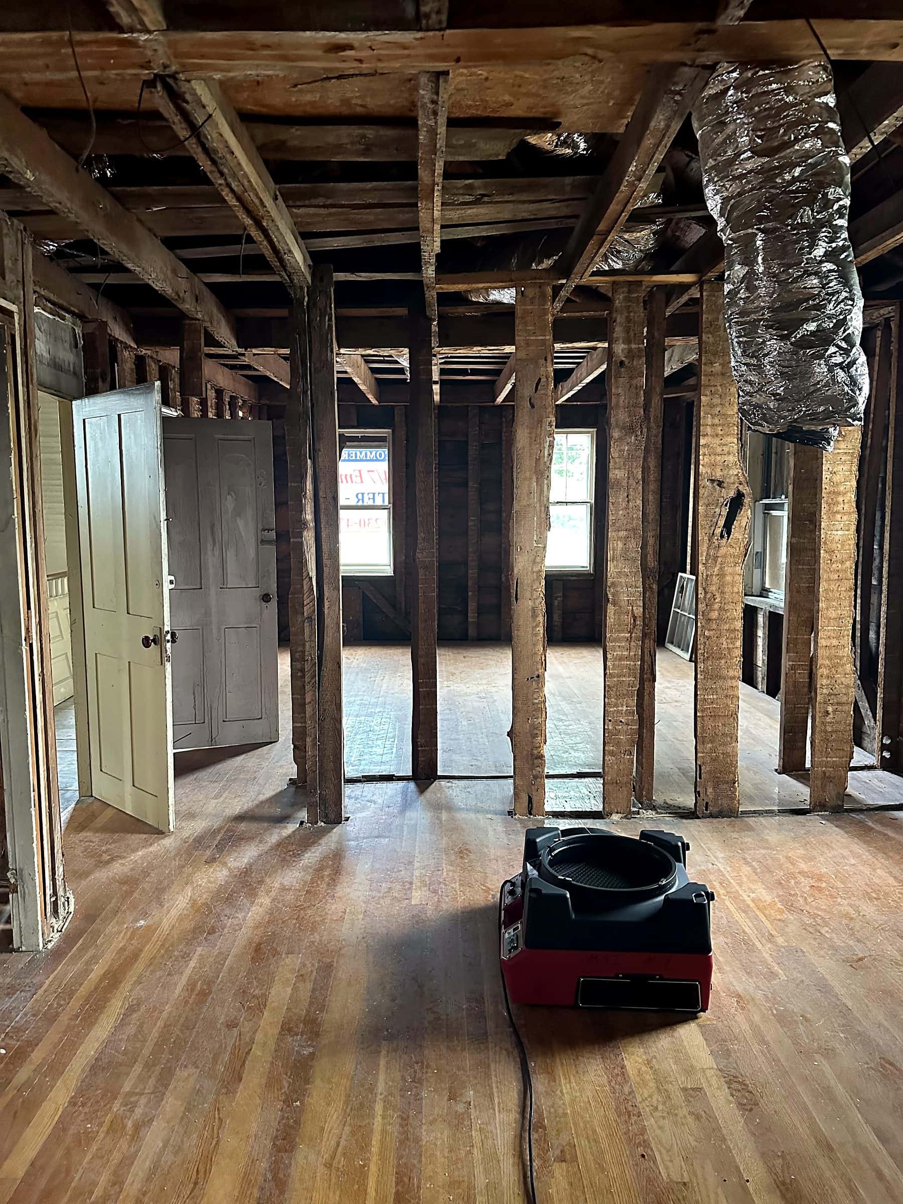 A room gutted to the framing with an air mover set on the hardwood floor.
