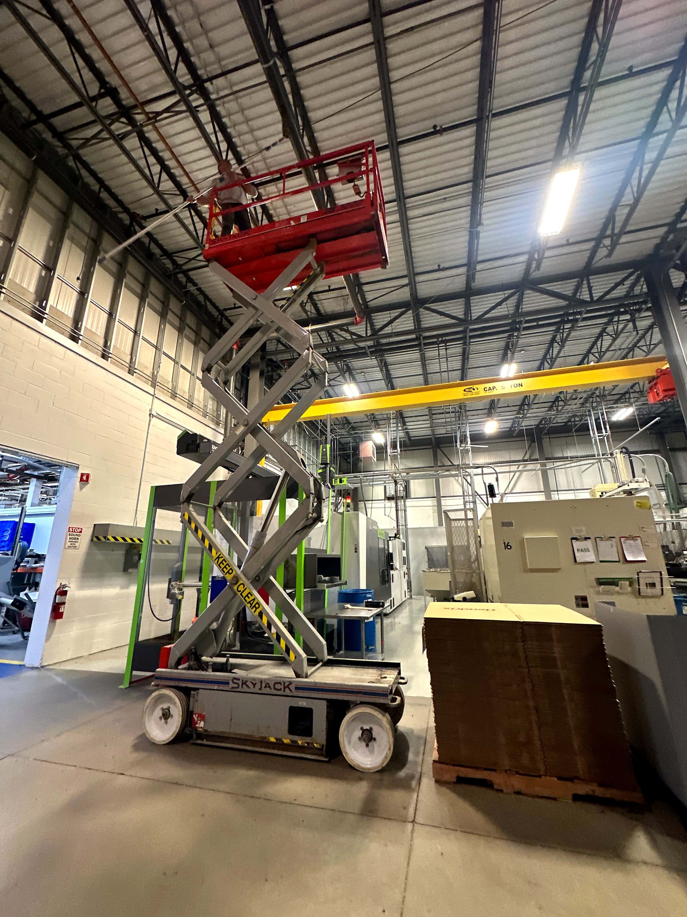 A scissor lift parked on the floor of a plant under high bay lights.